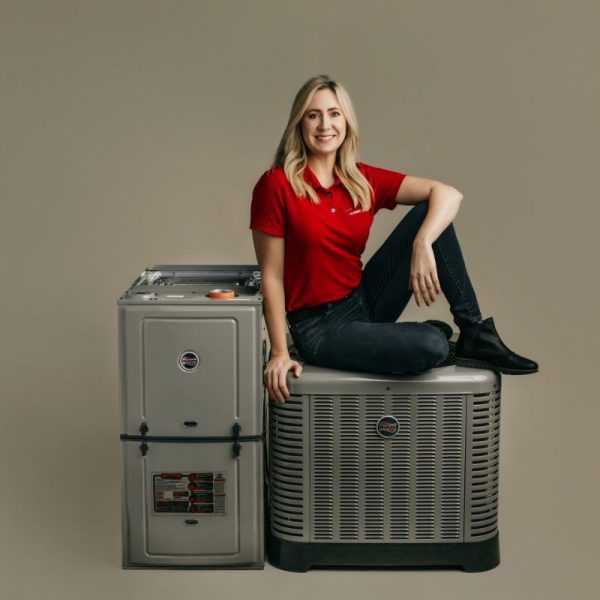 Holt-Amanda-On-Units A Person In A Red Shirt And Jeans Sits On An Air Conditioning Unit Beside A Furnace Against A Plain Background.